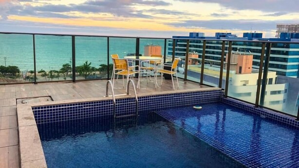 A rooftop pool with a view of the ocean, surrounded by glass railings and yellow chairs. - Grande Apartamento Beira-Mar (Maceió)