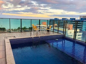 Piscina en la azotea con vista al océano, rodeada de barandillas de vidrio y sillas amarillas.