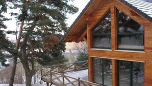 A wooden cabin with a balcony and large windows, surrounded by trees and a snowy landscape.