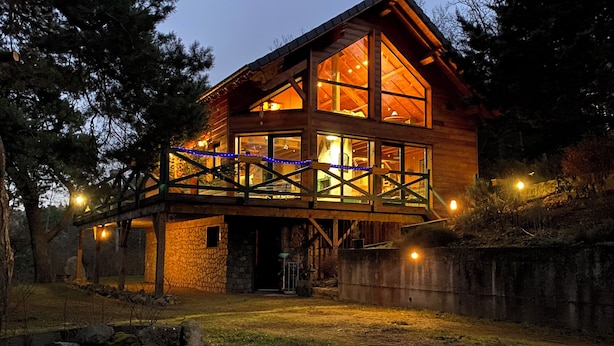 A wooden cabin with large windows, a stone foundation, and a wooden deck with string lights.