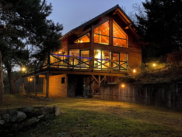 Una cabaña de madera con ventanas amplias, cimientos de piedra y una terraza de madera con luces colgantes.