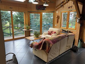 Interior acogedor de una cabaña con un sofá, una mesa y vistas a una terraza de madera y árboles.