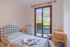 A bedroom with a checkered bedspread, a book, a wooden nightstand, a radiator, a wooden shelf, and a balcony with a view of houses and mountains.