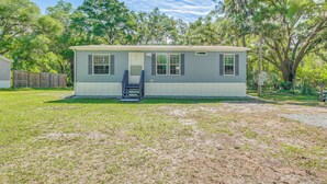 A single-story mobile home with a front porch and a small deck, surrounded by a grassy yard and trees. - 7 Mi to Crystal River: Home w/ Yard & Fire Pit! (Dunnellon)