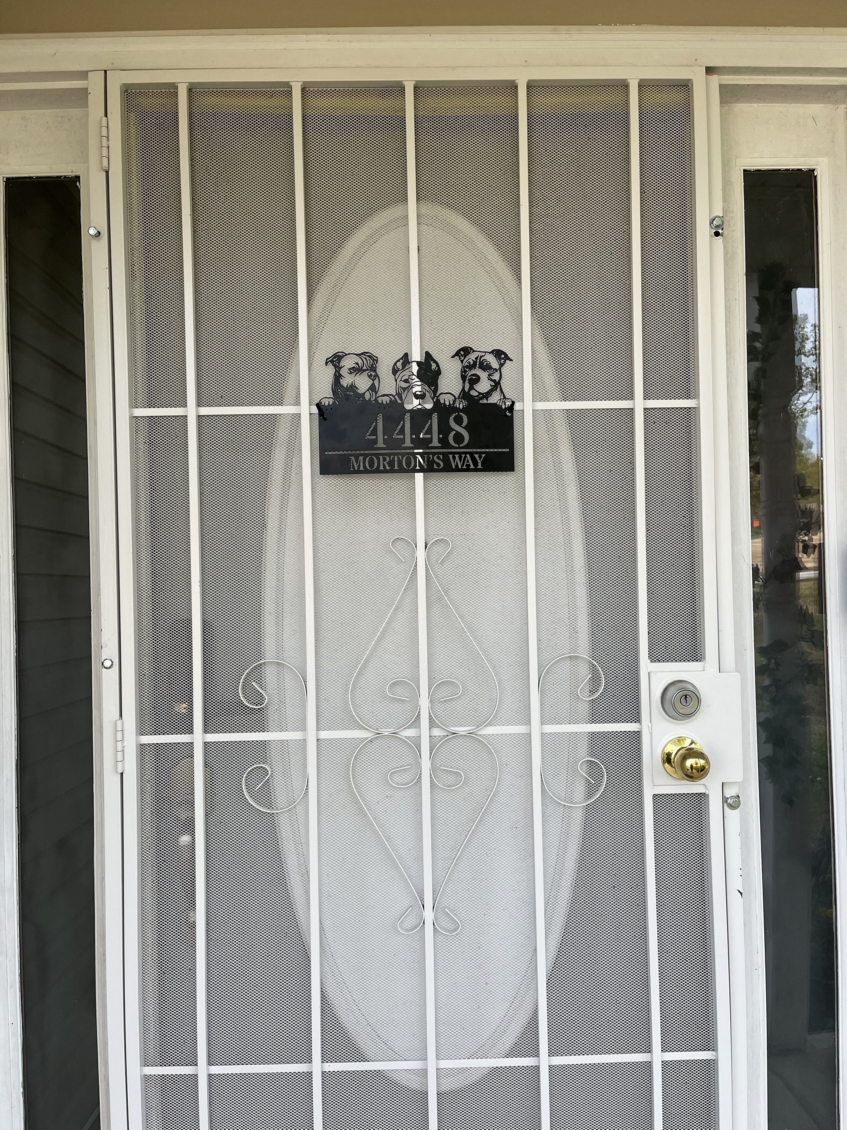 A house number sign with three dogs peeking over it, mounted on a white door with a metal grill.