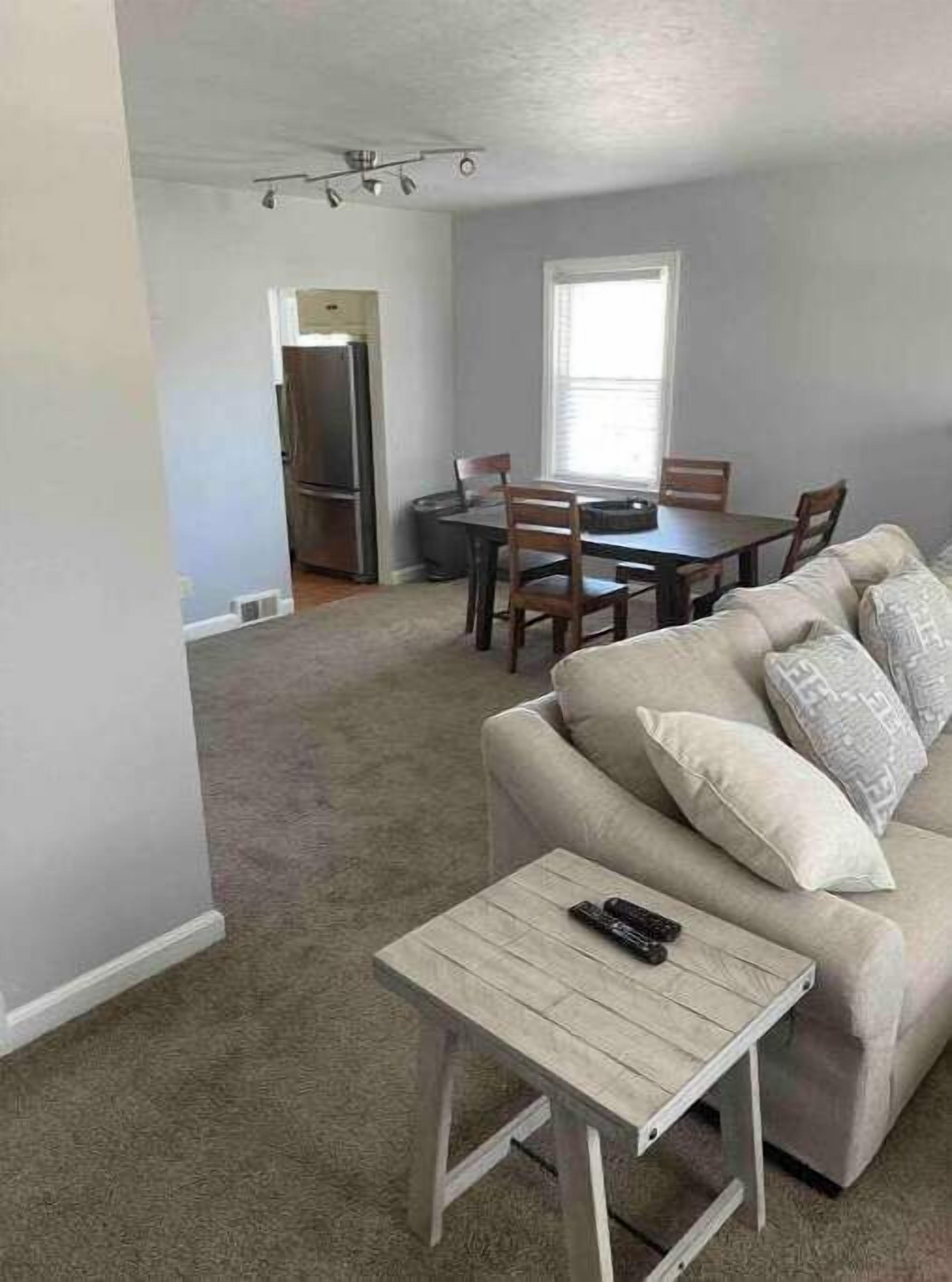 En stue med en beige sofa, et lille træsofa bord og et spiseområde med bord og stole.