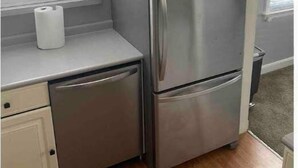 A compact kitchen with a stainless steel refrigerator, a dishwasher, and a window with blinds.