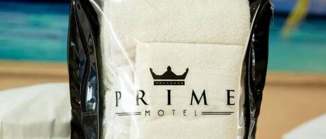 Una toalla blanca con el texto "PRIME MOTEL" y un logotipo de corona, sobre una cama, con un cielo azul y palmeras al fondo.
