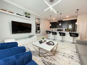 Una sala de estar moderna con un televisor de pantalla plana, sofás azules, una mesa de centro de mármol y una zona de cocina con taburetes de bar.