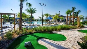 Zona de piscina con palmeras, sillas de descanso y un pequeño campo de práctica de golf.