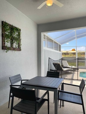 Un patio techado con mesa y sillas, un ventilador de techo, y vistas a una piscina y al paisaje.