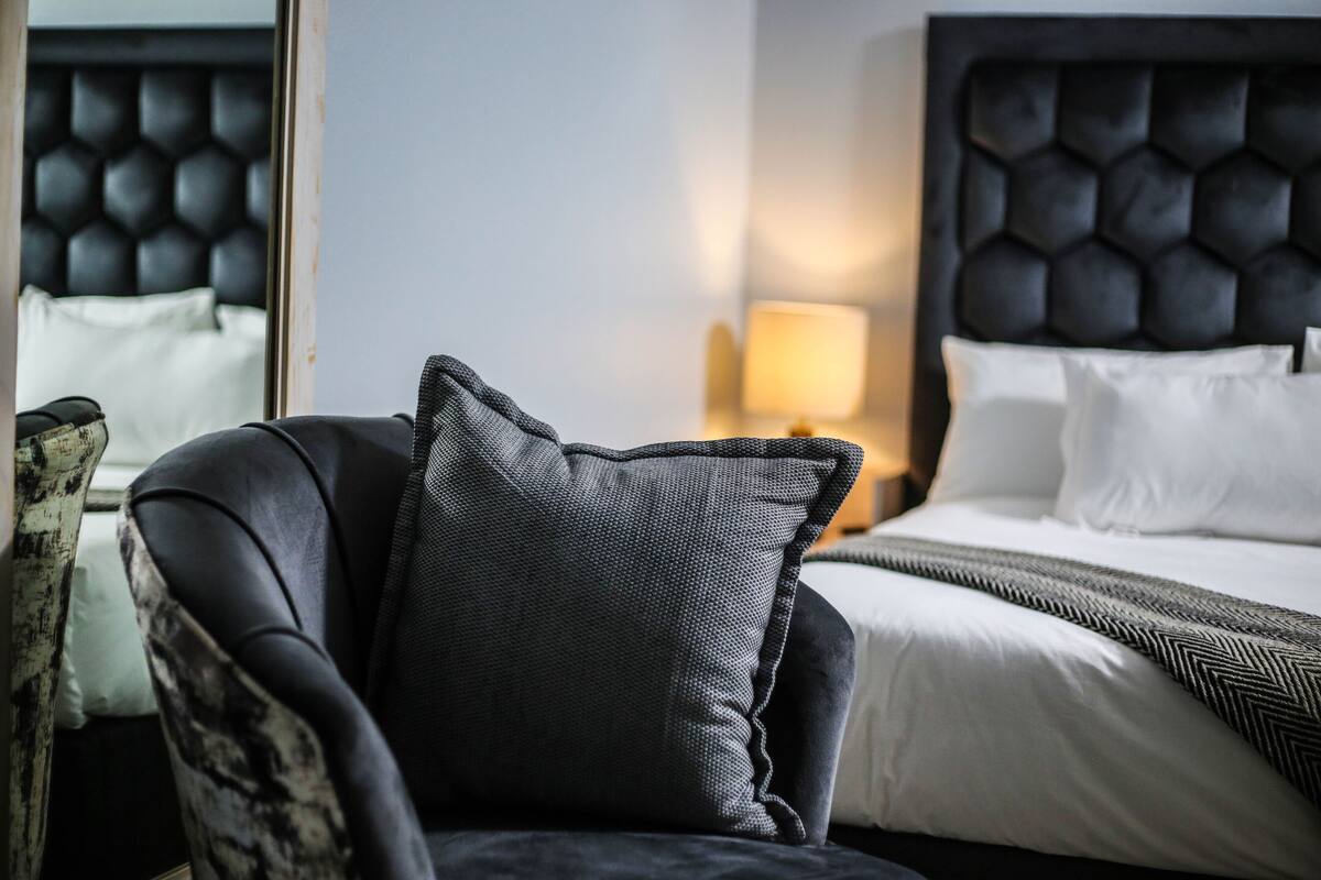 A modern hotel room with a tufted headboard, a dark armchair, and a neatly made bed with white linens.