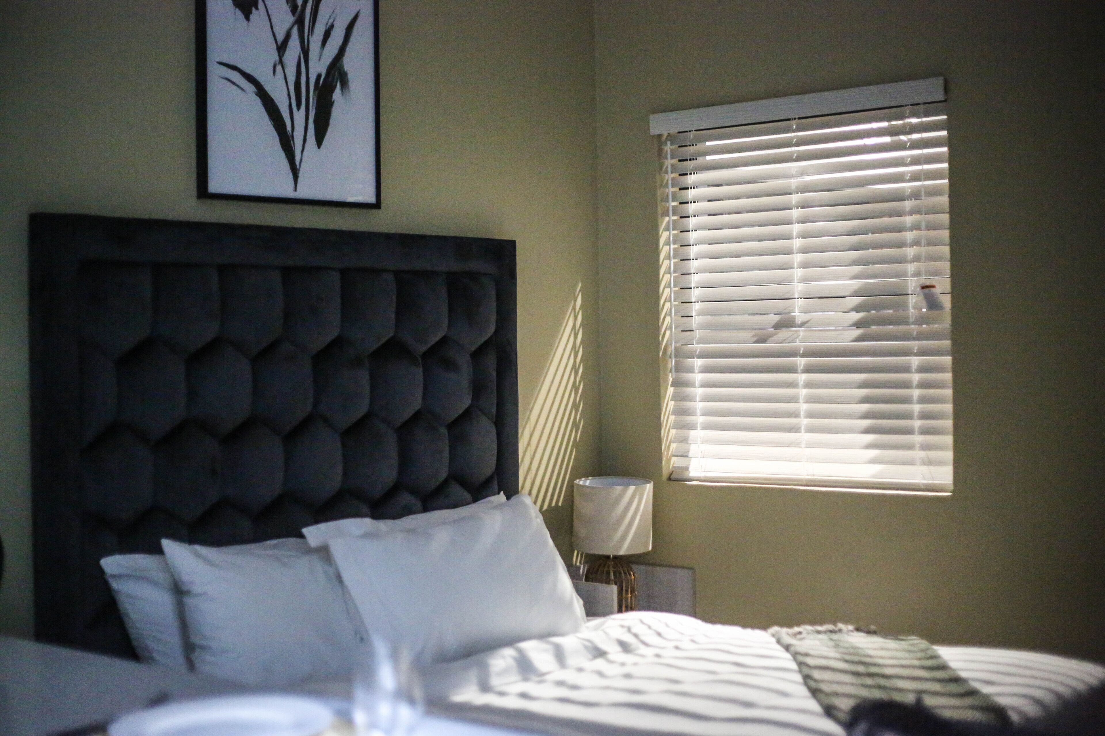 Un dormitorio con tablero de cama capitonado, cama con ropa de cama blanca, lámpara de noche y ventana con persianas.