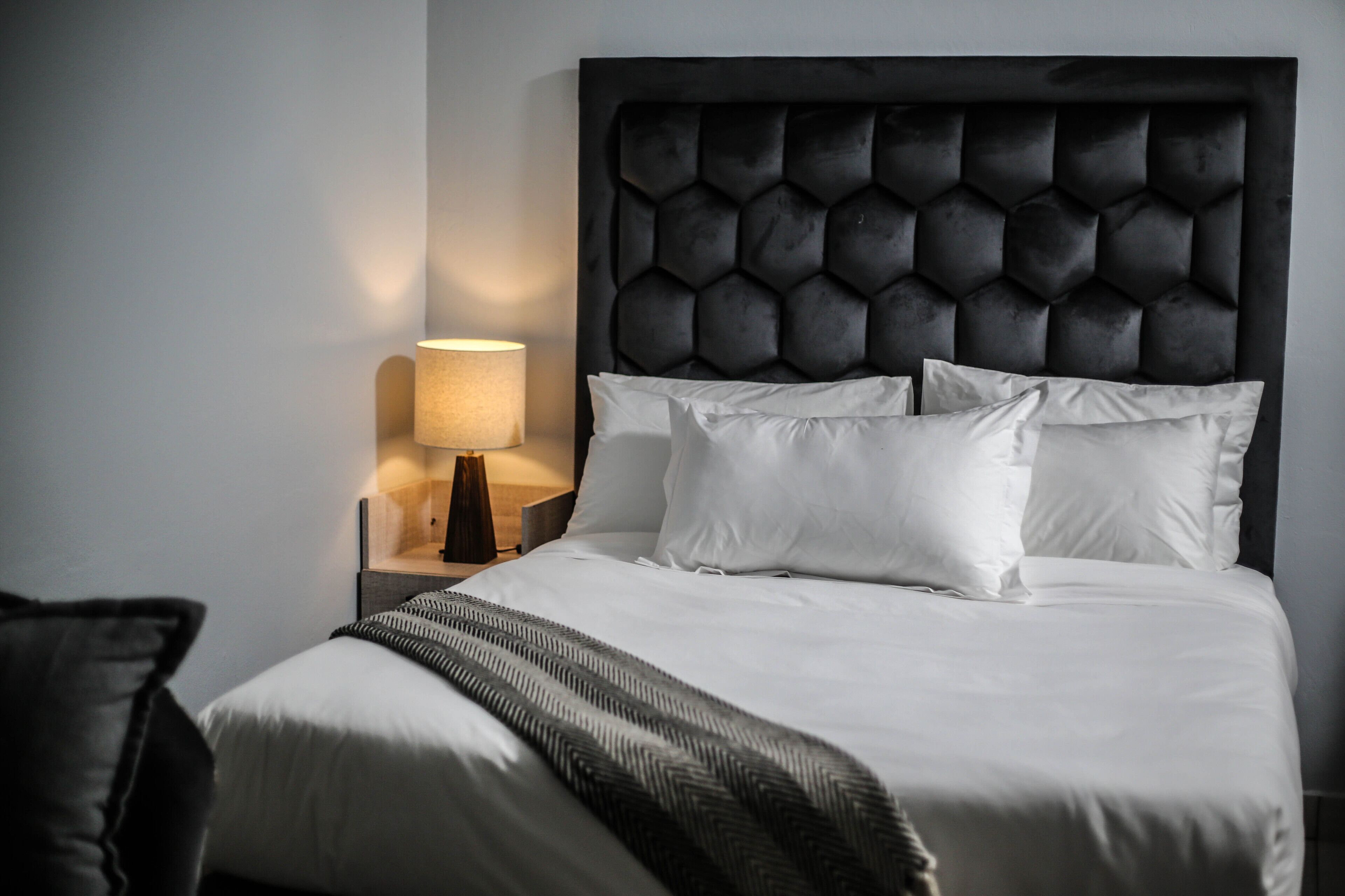 A neatly made bed with white linens, a dark headboard, and a bedside lamp.