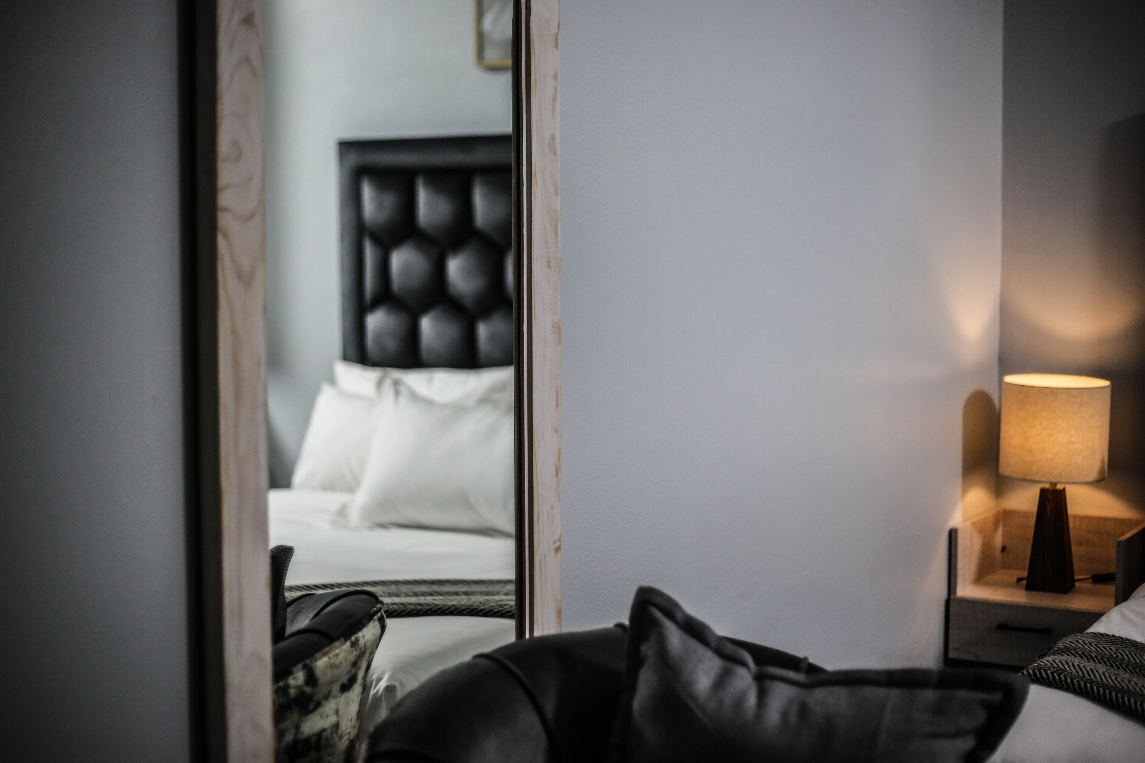 A hotel room with a bed, a nightstand with a lamp, and a mirror reflecting the room.