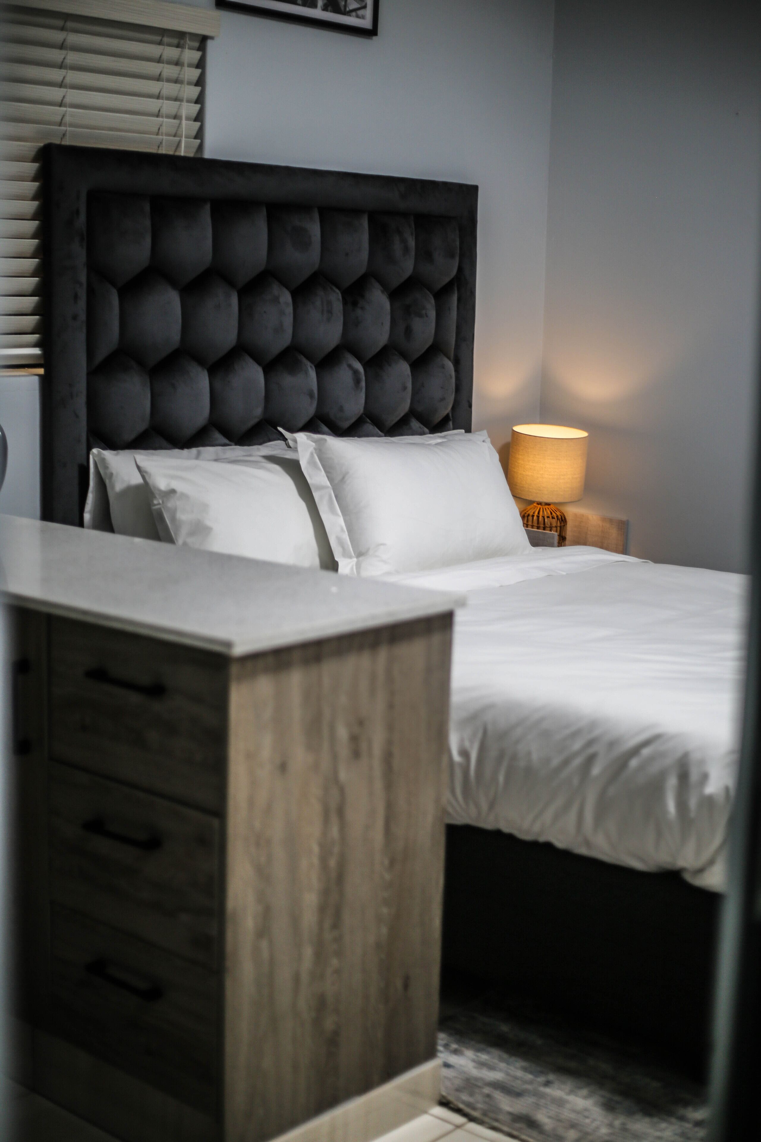 A bedroom with a tufted headboard, a bedside table, a lamp, and a bed with white bedding.