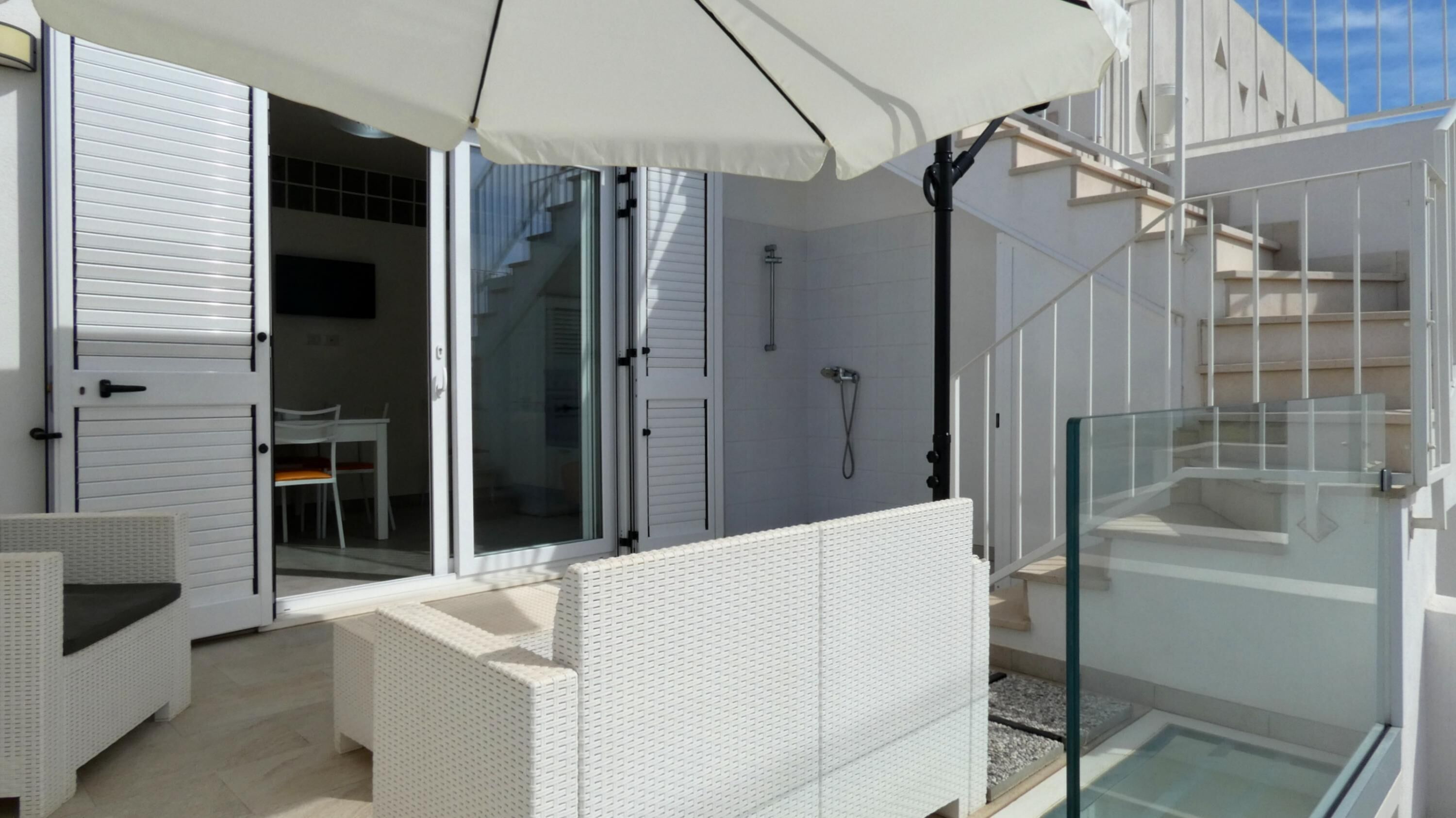 Un espacio exterior moderno con mobiliario de mimbre blanco, una ducha con cerramiento de vidrio y una escalera que lleva a un nivel superior.