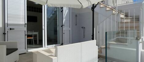 Un espacio exterior moderno con mobiliario de mimbre blanco, una ducha con cerramiento de vidrio y una escalera que lleva a un nivel superior.