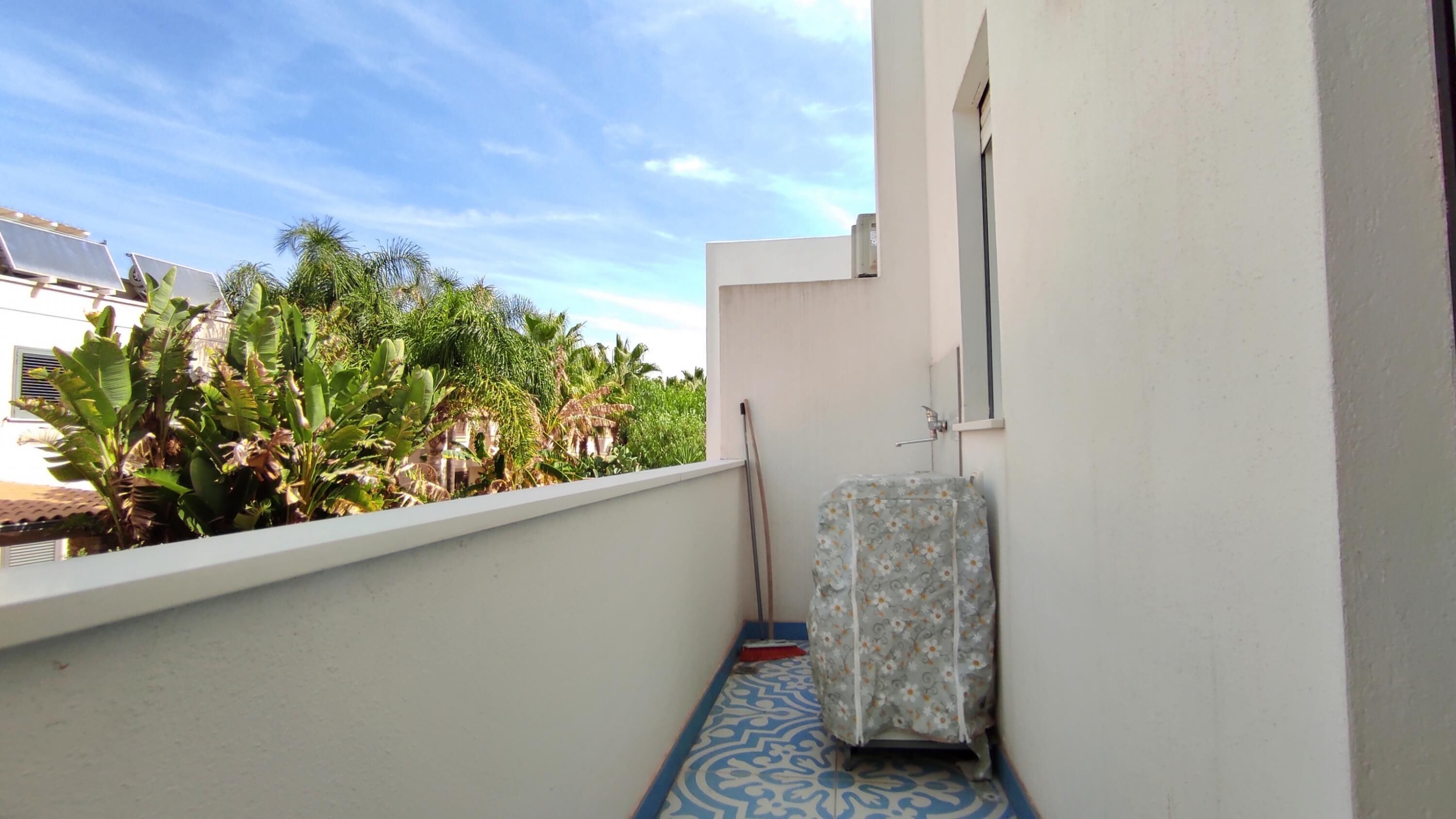 A narrow balcony with a patterned floor, a white wall, and a window overlooking a garden with various plants.