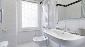 A modern bathroom with a white sink, bidet, and toilet. There is a window with blinds, a mirror, and a towel rack.