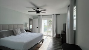 Ein Schlafzimmer mit einem Bett, einem Deckenventilator, einem Nachttisch mit Lampe, einem Fernseher und einem Fenster mit Vorhängen.