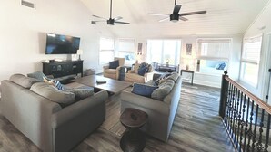 Ein geräumiges Wohnzimmer mit hoher Decke, einem Flachbildfernseher, mehreren Sitzgelegenheiten und großen Fenstern, die für natürliches Licht sorgen.
