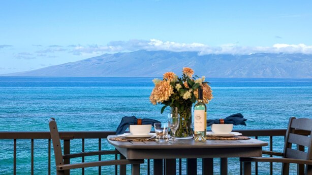 A table set with a vase of flowers, a bottle, and glasses, overlooking a body of water with mountains in the background. - Lokelani Resort by KBM (Napili-Honokowai)