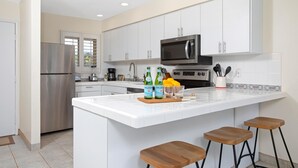 A modern kitchen with white cabinets, stainless steel appliances, a central island with stools, and a window with blinds. - Lokelani Resort by KBM (Napili-Honokowai)
