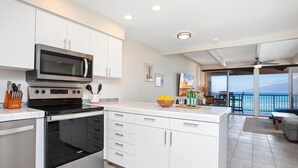 A modern kitchen with white cabinetry, a built-in microwave, and an oven. There is a dining area with a view of the ocean and a balcony. - Lokelani Resort by KBM (Napili-Honokowai)