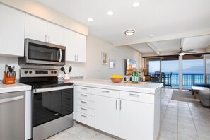 Una cocina moderna con armarios blancos, un microondas integrado y un horno. Hay un comedor con vistas al océano y un balcón.