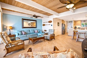 A spacious living room with a blue sofa, wicker furniture, a ceiling fan, and a kitchen area visible through an open door. - Lokelani Resort by KBM (Napili-Honokowai)