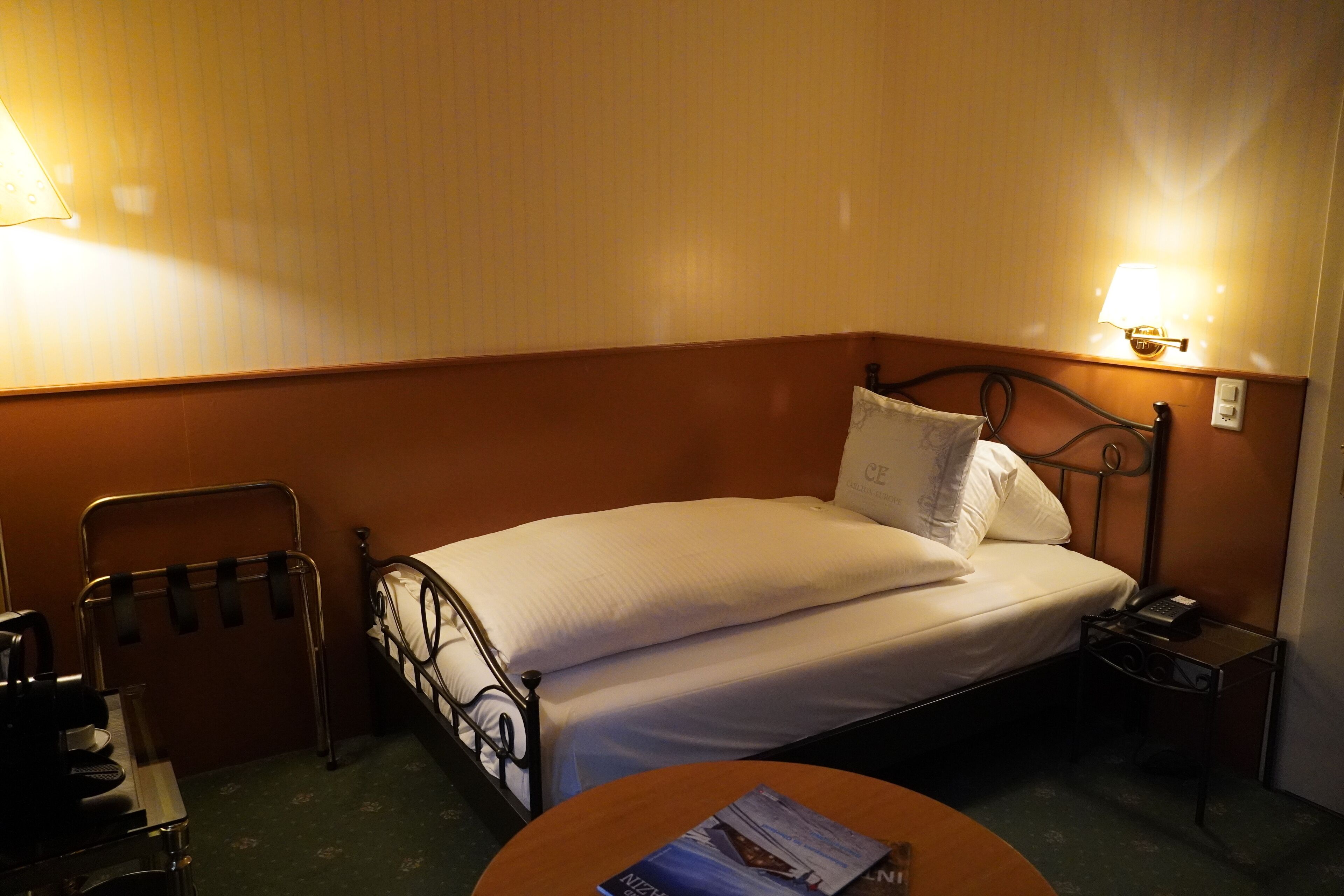 Quarto de hotel com uma cama de solteiro, mesa de cabeceira com abajur, uma cadeira e uma pequena mesa com uma revista.
