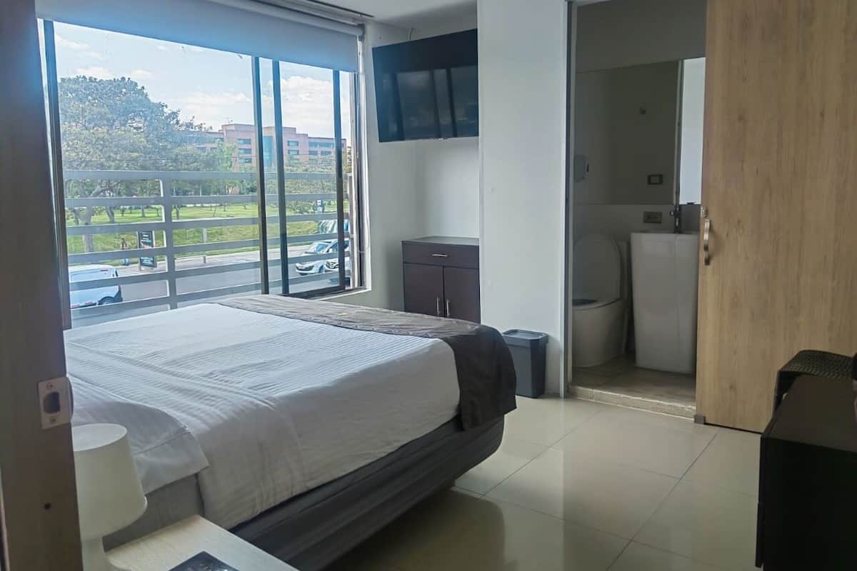 Ein modernes Hotelzimmer mit einem großen Bett, einem an der Wand montierten Fernseher, einer kleinen Kommode und Blick ins Freie durch ein großes Fenster.