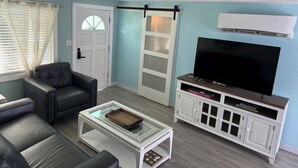 A living room with a leather sofa, a glass coffee table, a TV on a wooden stand, and a white cabinet. - Northern Point of Treasure Island, NPR - waterfront w/ pool, sunroom & kayaks! (New Port Richey)