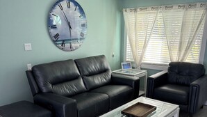 A living room with a black leather sofa, a glass coffee table with a tray, and a large wall clock. - Northern Point of Treasure Island, NPR - waterfront w/ pool, sunroom & kayaks! (New Port Richey)