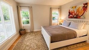 A bedroom with a bed, a window with blinds, a bedside table with a lamp, a wall with a floral painting, and a rug on the floor.