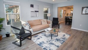 Un salon spacieux comprenant un canapé, une table basse et un fauteuil. On y trouve également un ventilateur de plafond et une fenêtre avec des stores.