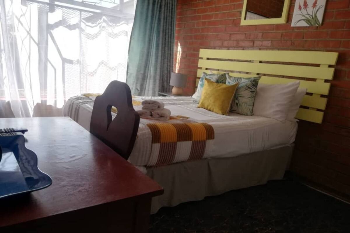 A hotel room with a bed, a desk, and a chair. There is a window with curtains, a lamp on the bedside table, and a picture on the wall.