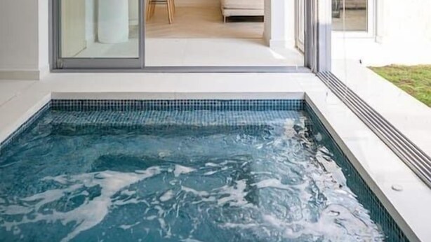 A modern indoor swimming pool with glass walls, a seating area with a sofa and chairs, and a decorative wall.