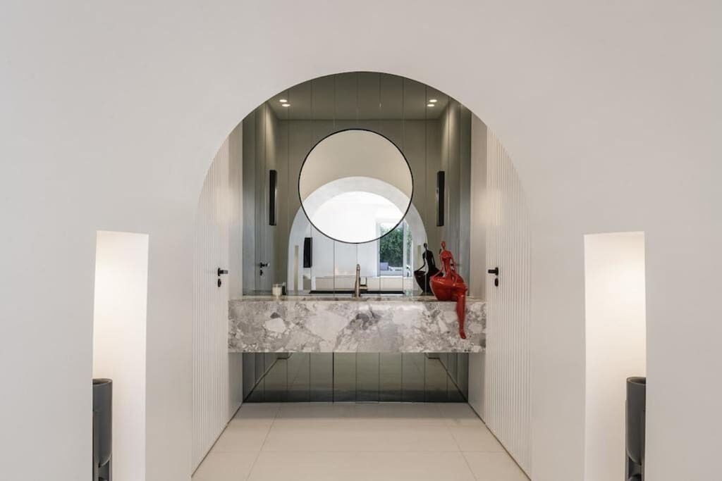 Une salle de bain moderne avec un grand miroir, un comptoir en marbre et un objet décoratif rouge.