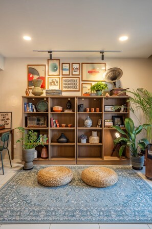 Une pièce comprenant une bibliothèque en bois, deux poufs en fibres tressées, un tapis bleu à motifs et divers objets décoratifs.