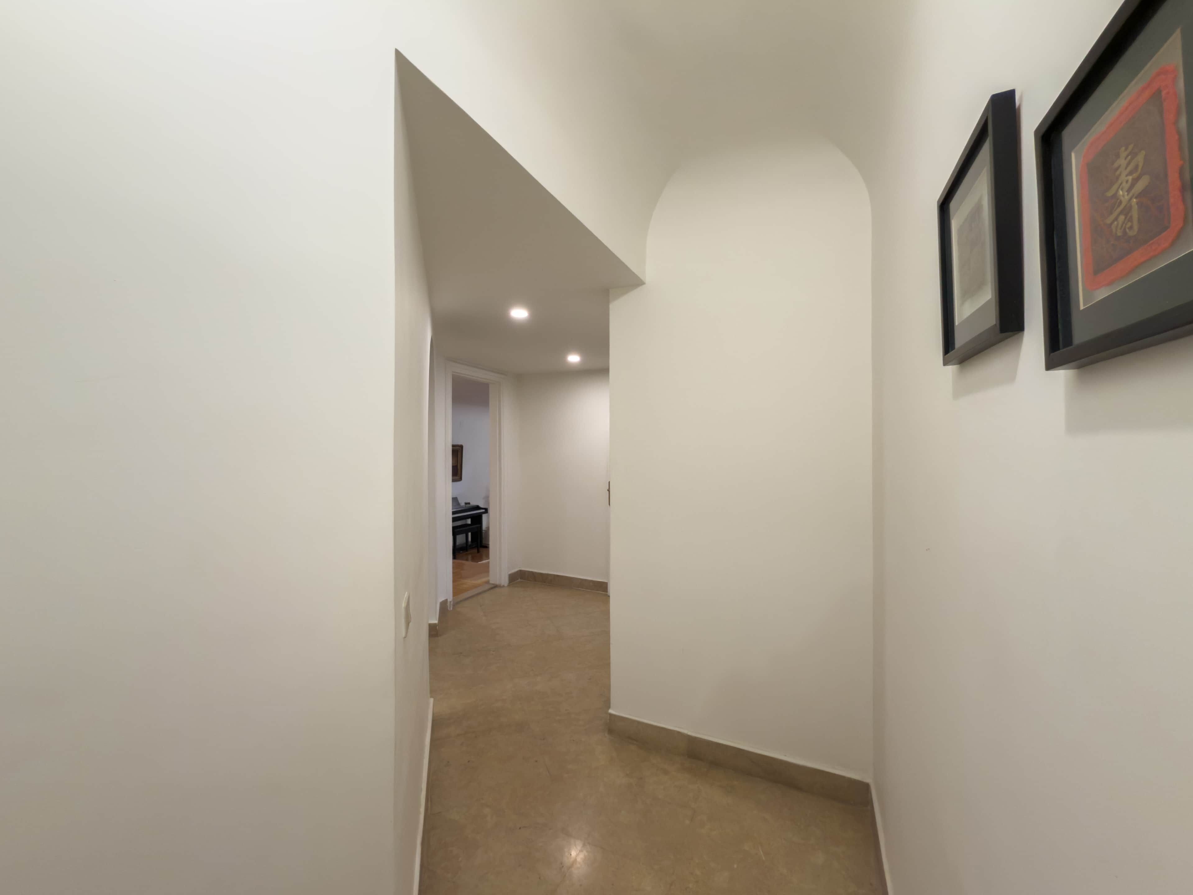 A hallway with white walls and a brown floor, leading to a room with a desk and chair.