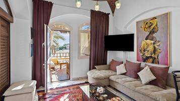 A living room with a sofa, a coffee table, and a TV. There is a painting on the wall and a window with curtains.