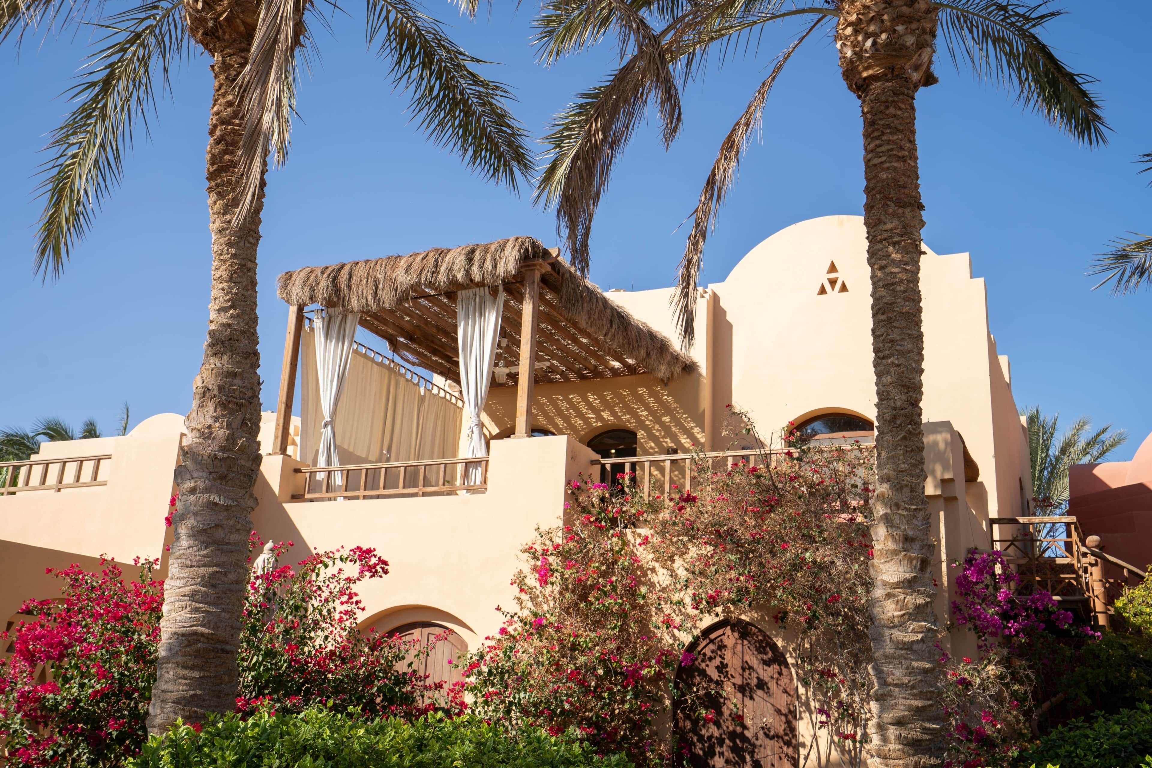 A building with a thatched roof balcony, palm trees, and pink bougainvillea.