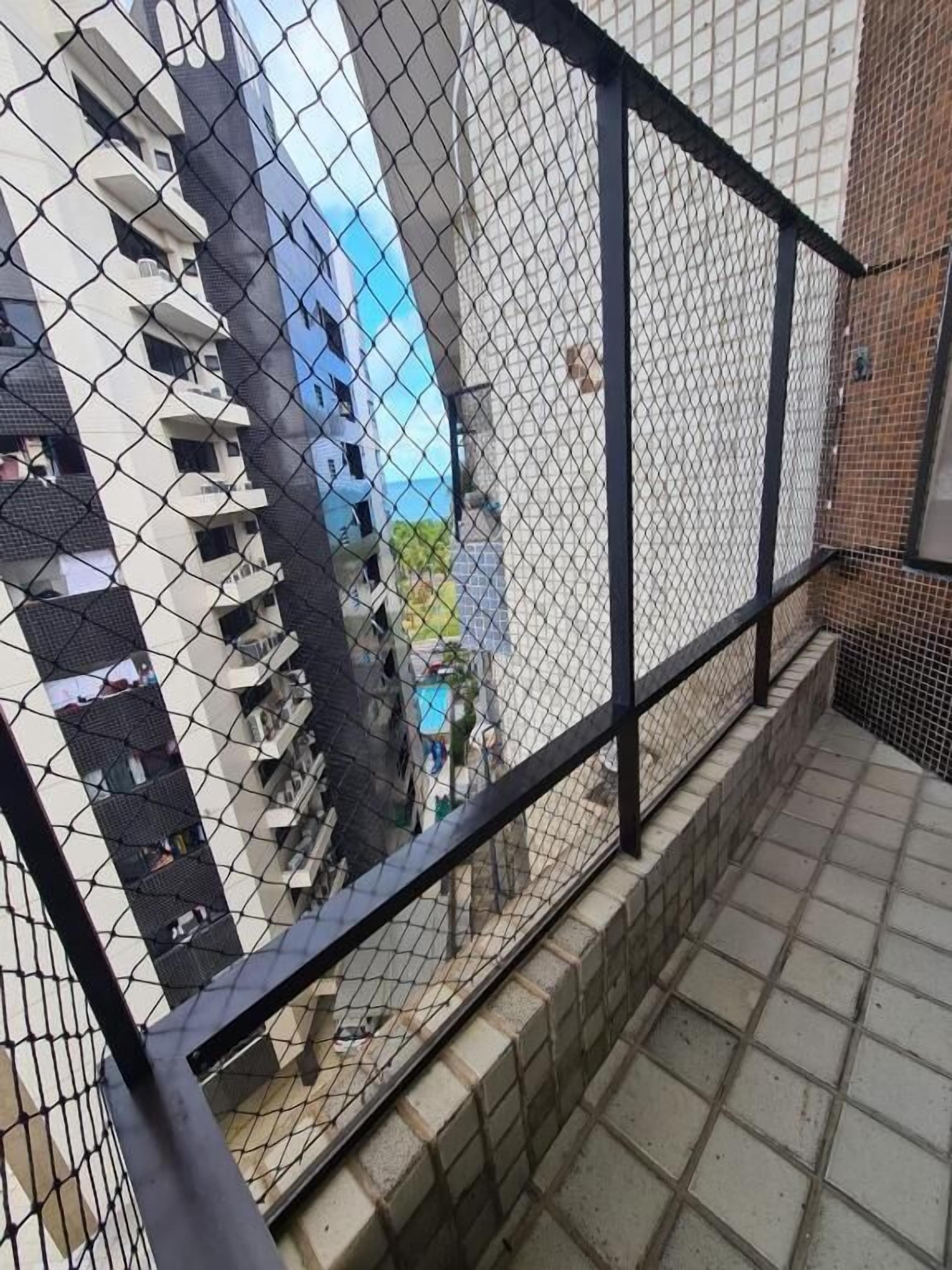 Una cerca de malla con baranda metálica negra, suelo de baldosas y vista a un edificio con varios balcones.