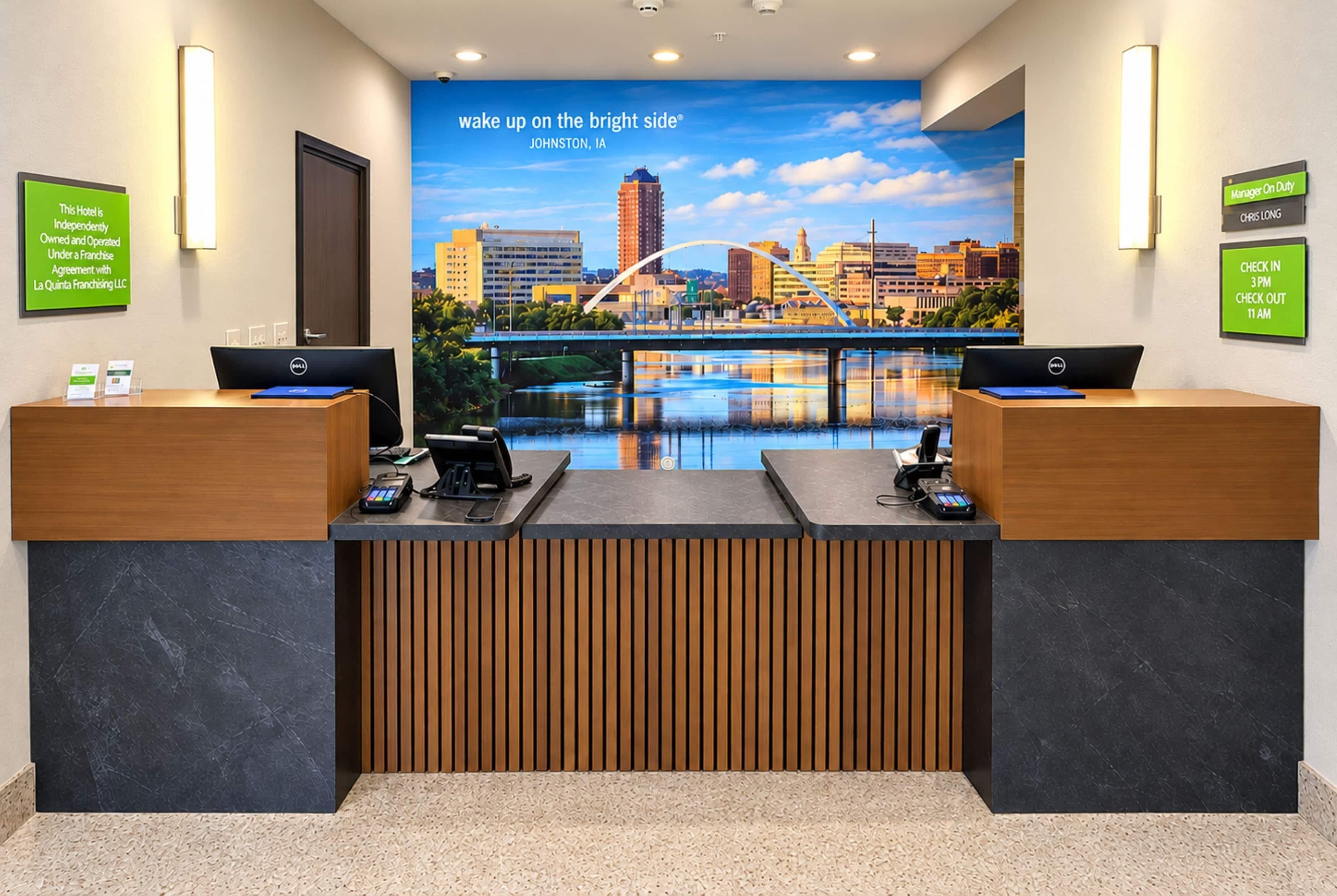 A hotel reception desk with two computer stations, a cityscape mural, and signage for check-in and check-out times.