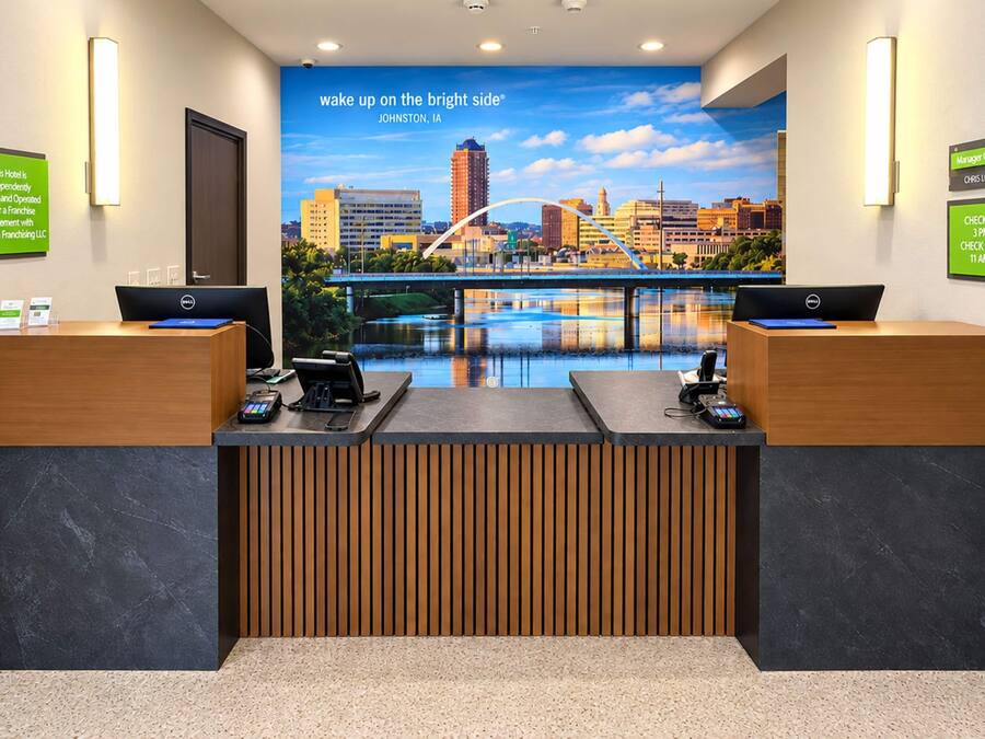 Ein Hotelrezeptionstresen mit zwei Computerarbeitsplätzen, einer Stadtszenen-Mural und Schildern für Check-in- und Check-out-Zeiten.