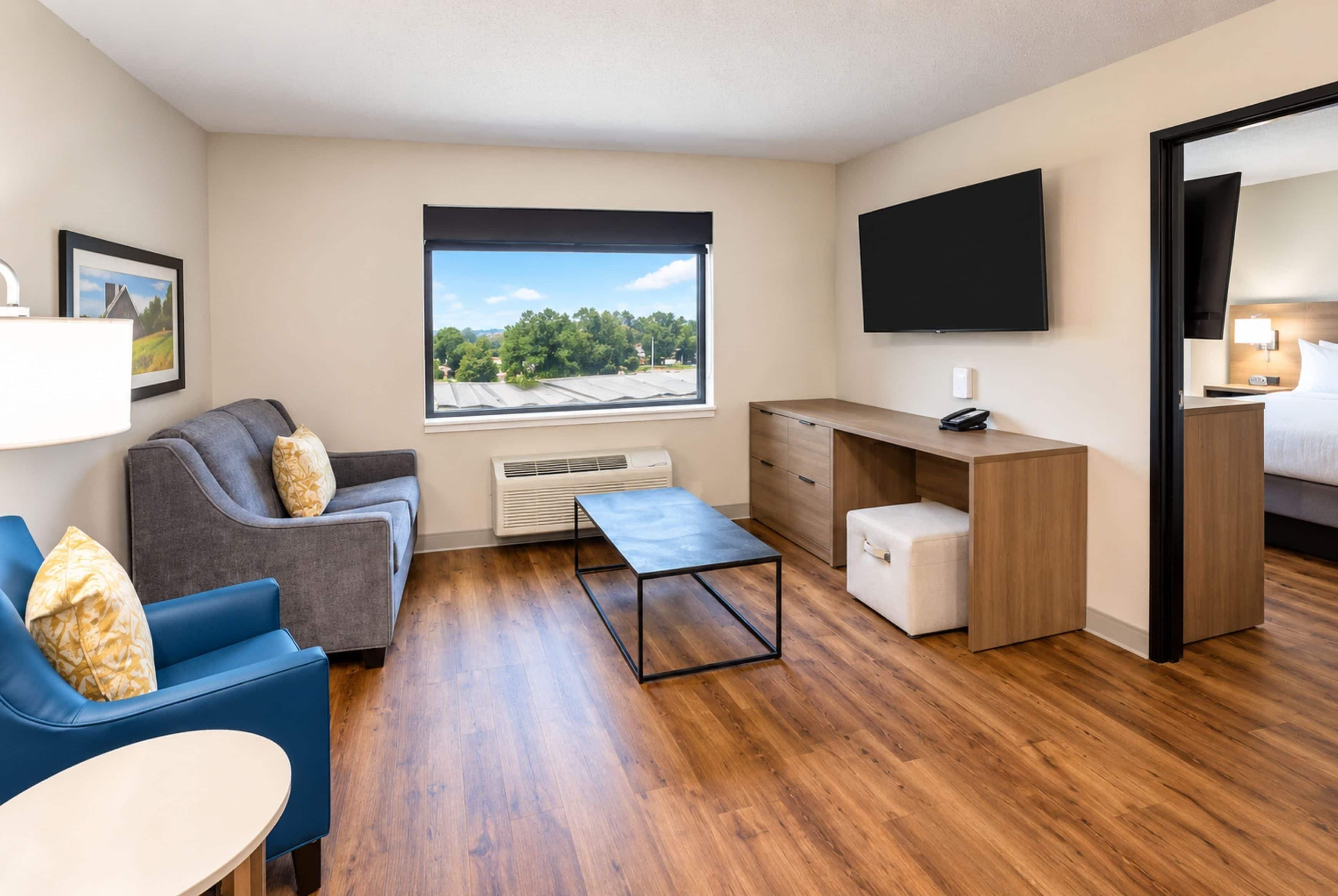 Una habitación moderna y bien iluminada con un sofá gris, una silla azul, un escritorio de madera y un televisor fijado a la pared.