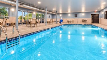 An indoor swimming pool with clear blue water, surrounded by lounge chairs and tables, and large windows offering views of greenery outside.