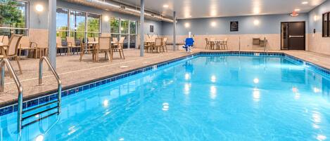 An indoor swimming pool with clear blue water, surrounded by lounge chairs and tables, and large windows offering views of greenery outside.